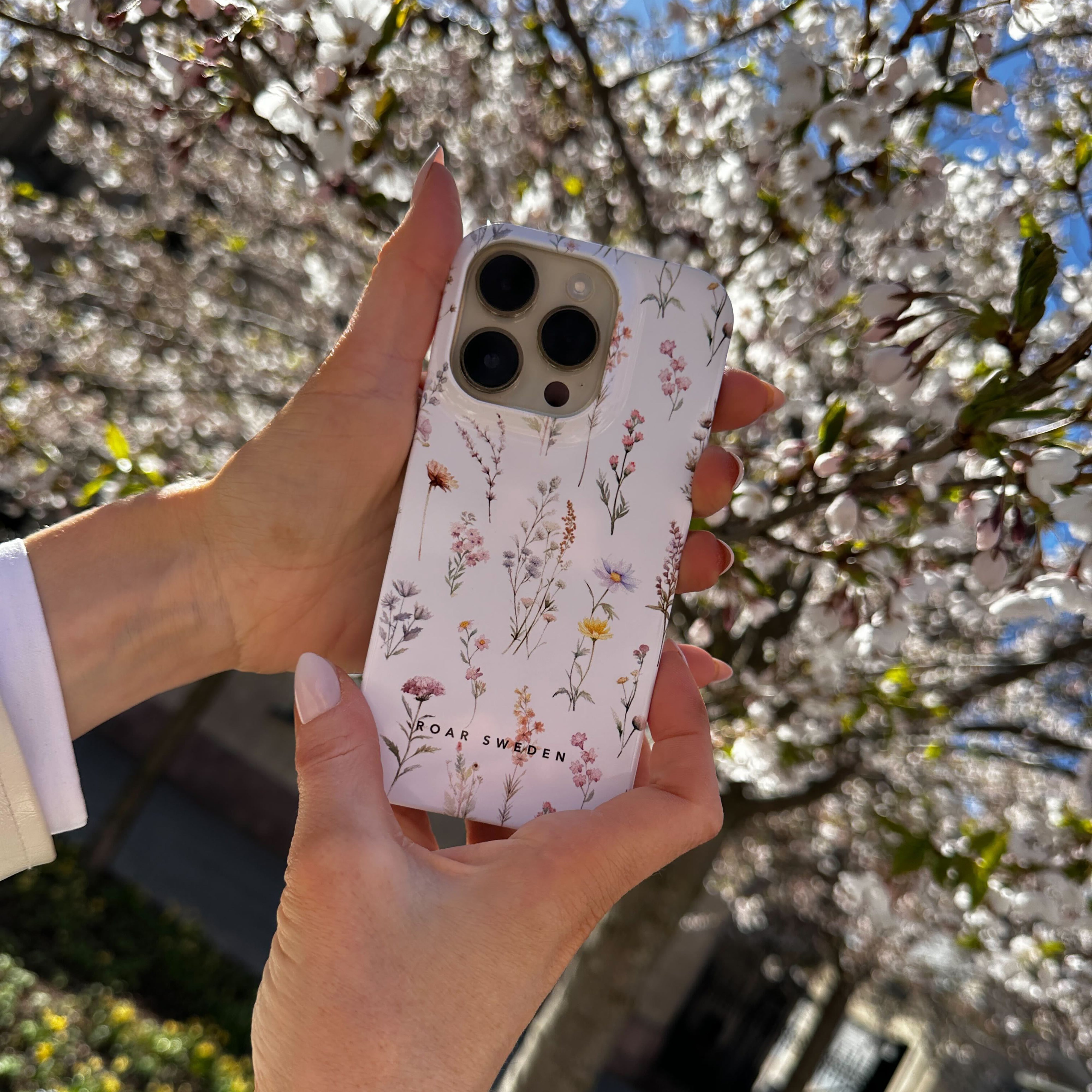 A person holds a Wild Flowers - Slim case-covered smartphone in front of blooming cherry blossom trees, reminiscent of a skandinavisk flora scene.