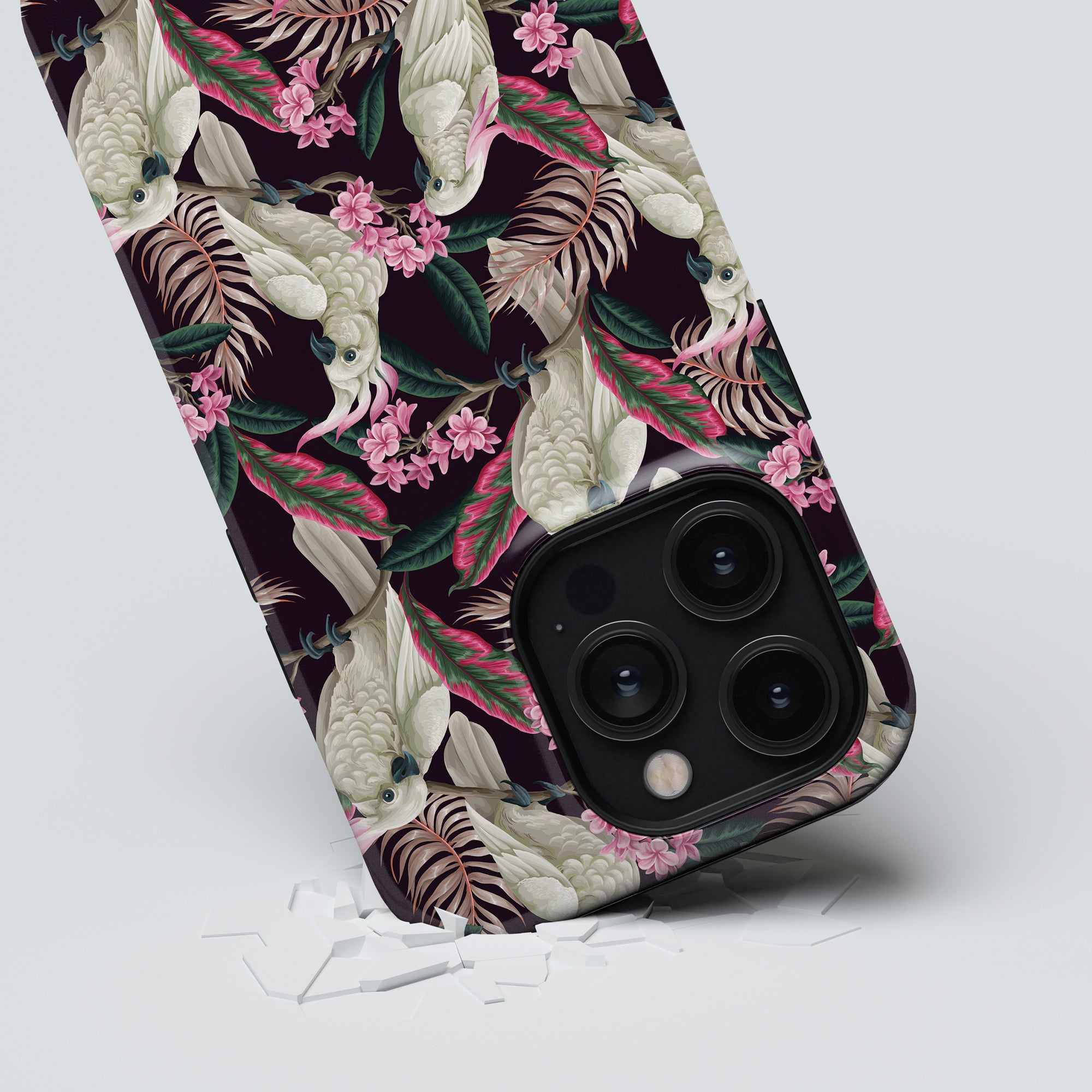 A smartphone with a tropical bird and floral patterned Cockatoo - Tough case is propped up, with a small amount of shattered glass beneath it.