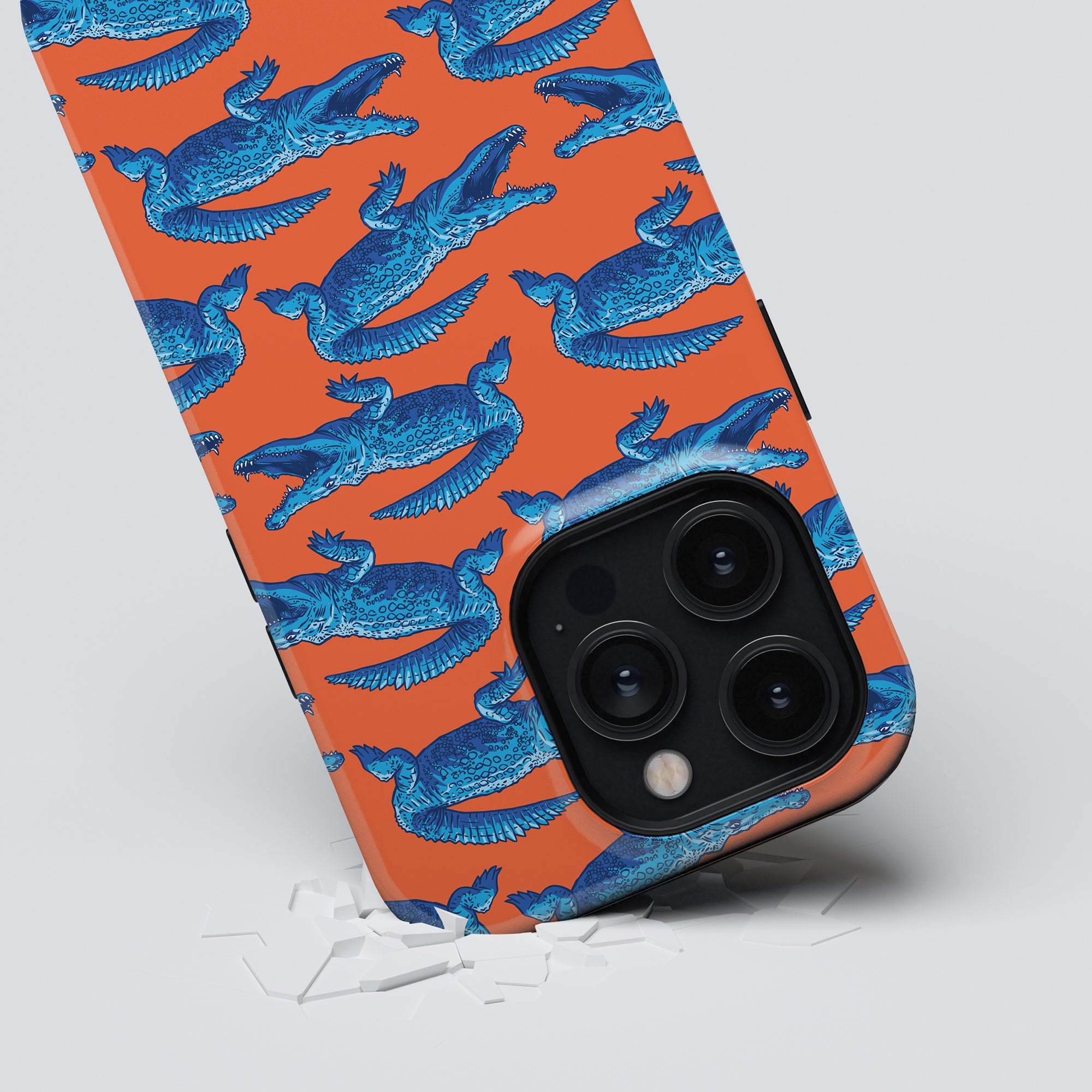 A smartphone in a Groovy Croc - Tough case from Roar Sweden, showcasing blue bird patterns on an orange background, lies tilted on a white surface with small shards underneath. The prominent rear camera module adds a touch of elegance to the design.