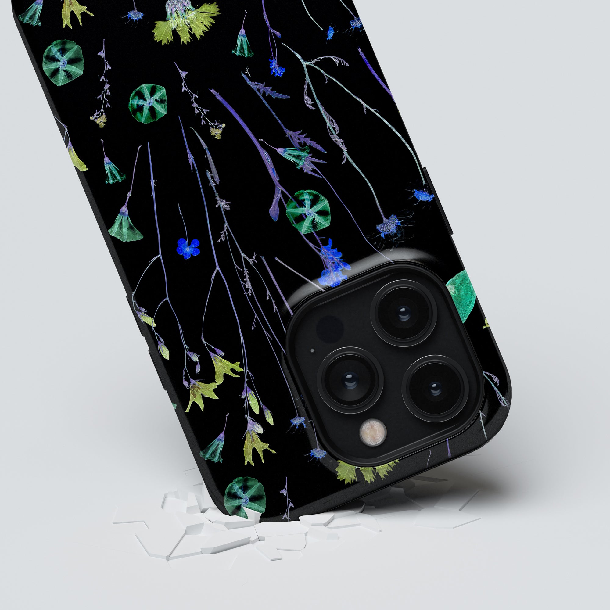 A smartphone enclosed in the Night Plants - Tough case, adorned with a floral pattern, lies on a white surface next to small shattered fragments. The phone's camera lenses are clearly visible, highlighting both style and robust protection.