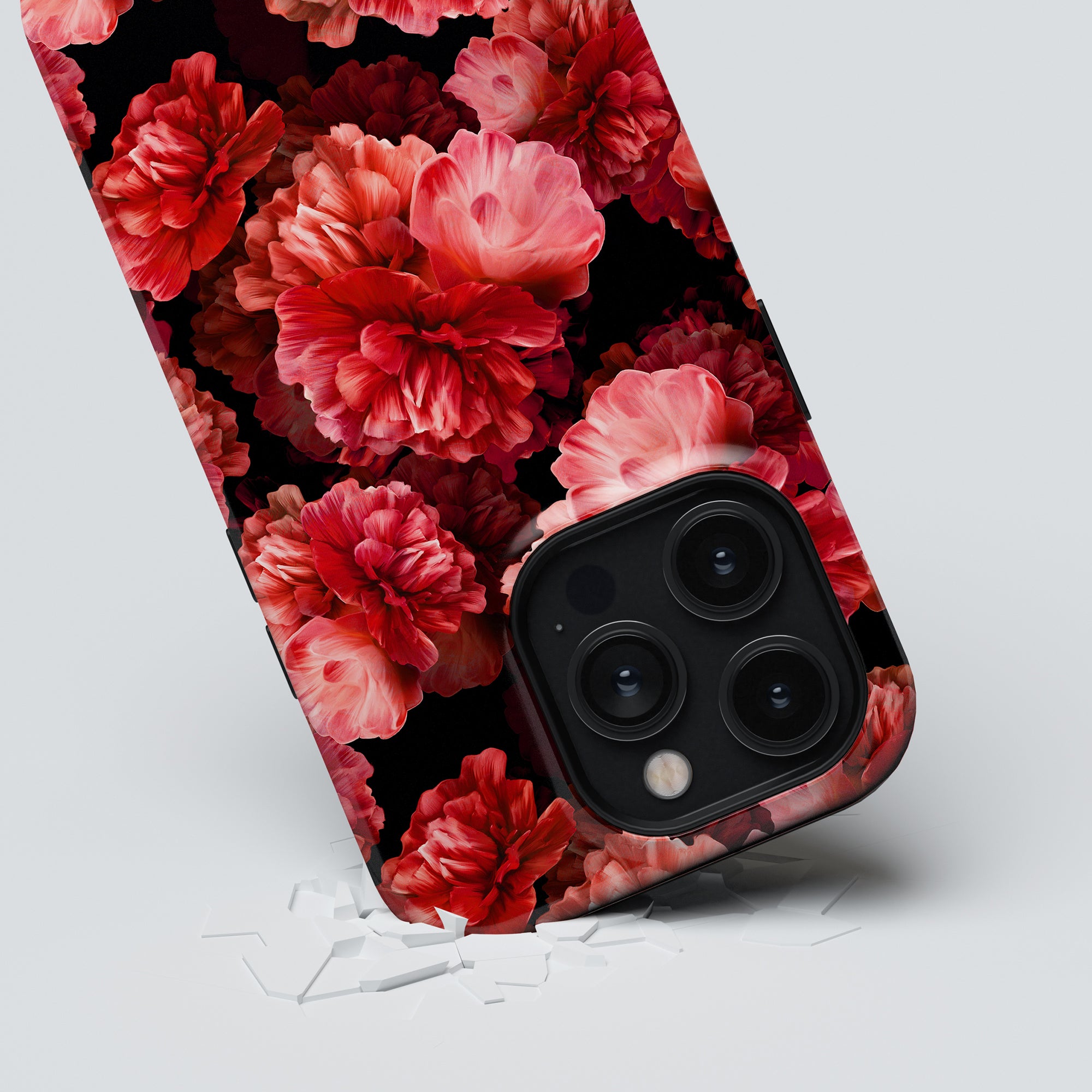 A smartphone with a triple-lens camera lies face-down on a cracked surface. The Peonies - Tough case showcases the blomstrande skönhet of red and pink flowers against a black background, providing not only style but also förstklassigt skydd.