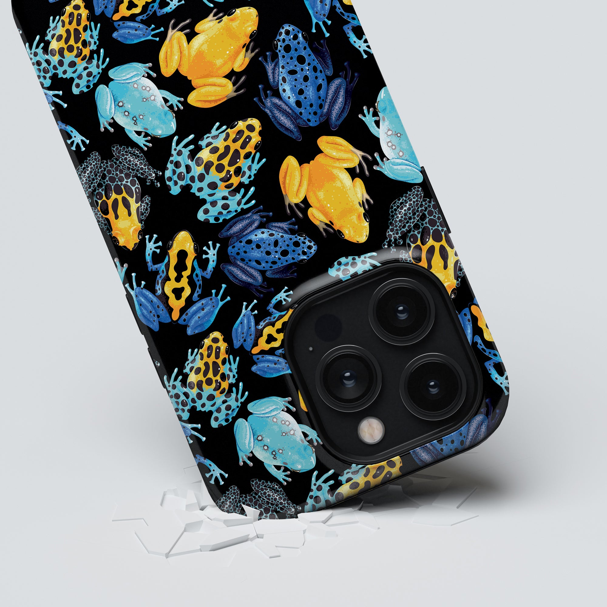 A smartphone with a vibrant Tropical Frogs - Tough case rests at an angle, with fragments of a shattered screen protector underneath.